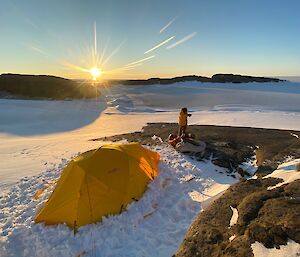 A yellow tent set up in the snow with rocks around.  An expeditioner stands to the side.