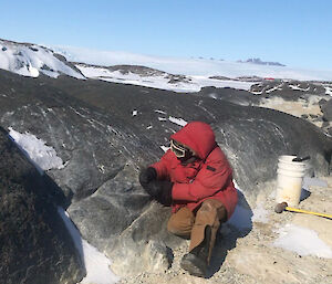 Expeditioner crouches by some rocks with buckets in the background