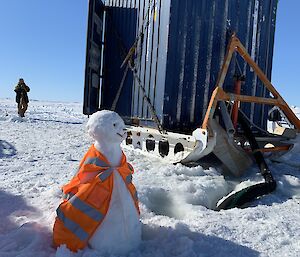 A snowman wearing hi-vis in front of a metal shed on a sled