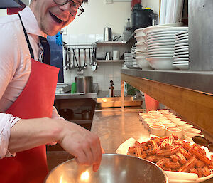 Expeditioner standing up in the kitchen sprinkling churros with sugar and smiling at the camera