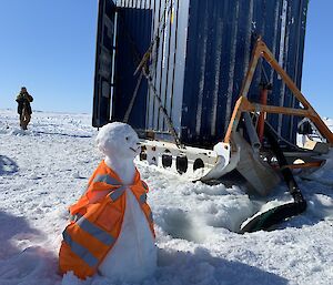 A snowman in an orange high vis jacket beside a seawater intake pipe, with a shipping container behind.