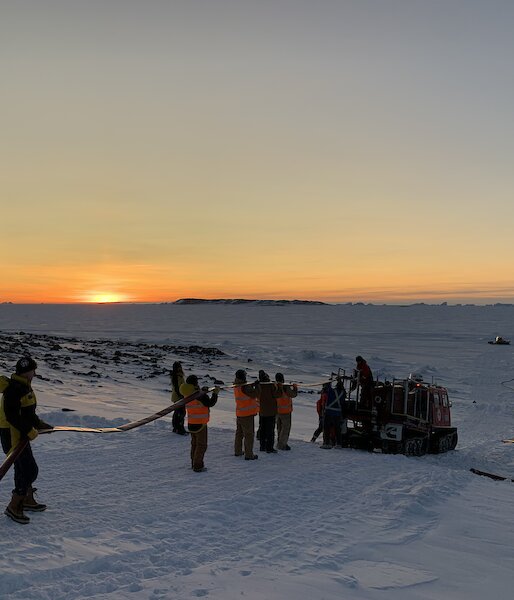 People in a line holding a hose being fed into a hagglund on the ice, with a sunset on the horizon.