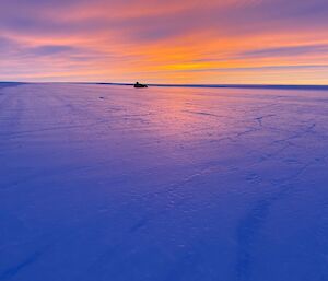 A proof roller in the distance at sunset with purple blue orange hues in the sky and on the ice