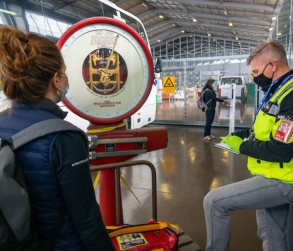 Two people wearing masks weigh a red bag on a scale, beside a bus, while another person uses a hand sanitizer dispenser in the background.