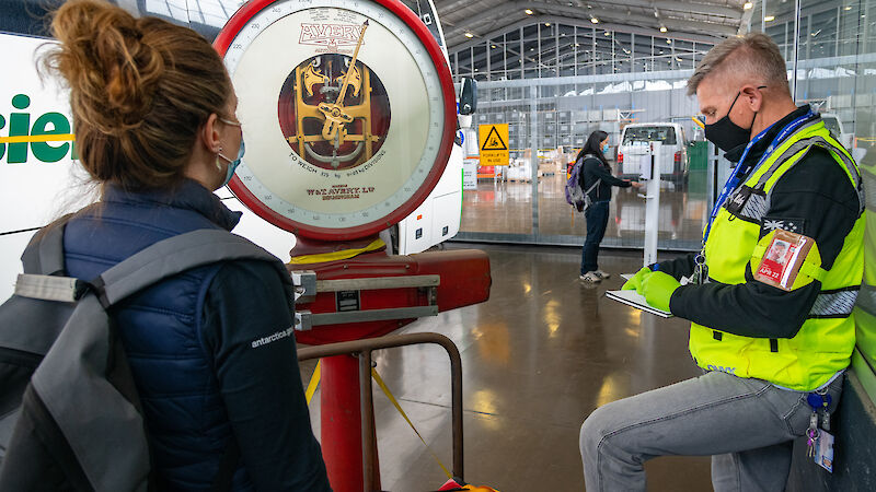 Two people wearing masks weigh a red bag on a scale, beside a bus, while another person uses a hand sanitizer dispenser in the background.