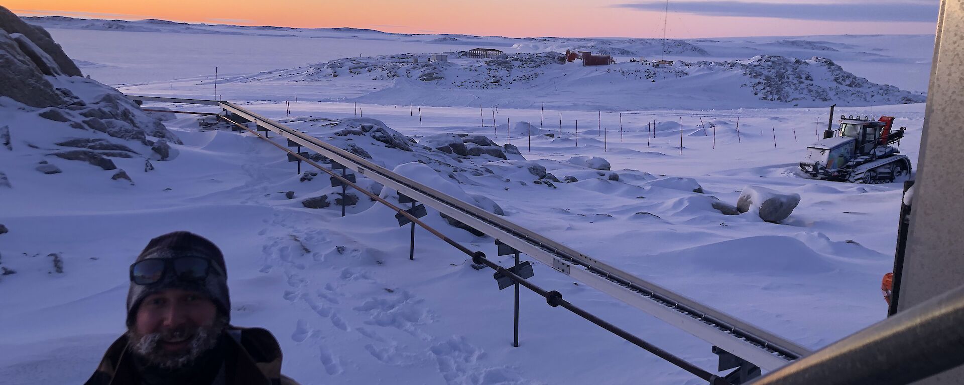 Expeditioner walking up stairs in foreground with fuel pipes behind him against a snowy landscape