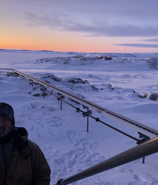 Expeditioner walking up stairs in foreground with fuel pipes behind him against a snowy landscape