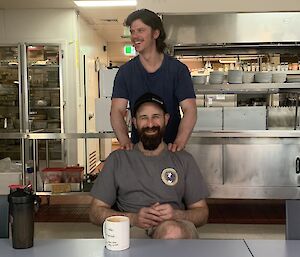 One expeditioner seated at the kitchen bench with another behind giving him a shoulder massage