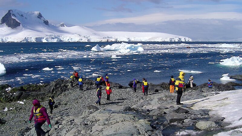 people in colourful jackets walk on rocky shore near icebergs