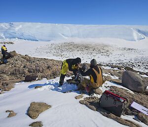 Three expeditioners sit atop a hill looking down on to Taylor penguin rookery.  Two expeditoners are taking photos of the penguins for research purposes.