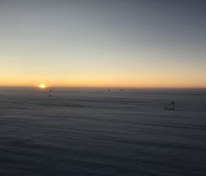 A landscape of the Wilkins Aerodrome with aeroplane stairs and a wind sock just visible in the blowing snow.  A sun is just rising on the horizon