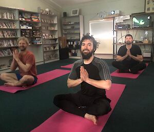 Three expeditioners sitting cross legged on yoga mats, with palms pressed together, looking to camera