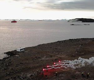 Expeditioners stand on a shoreline holding bright red flares with smoke plumes.  The Aurora Australis ship sail towards the horizon