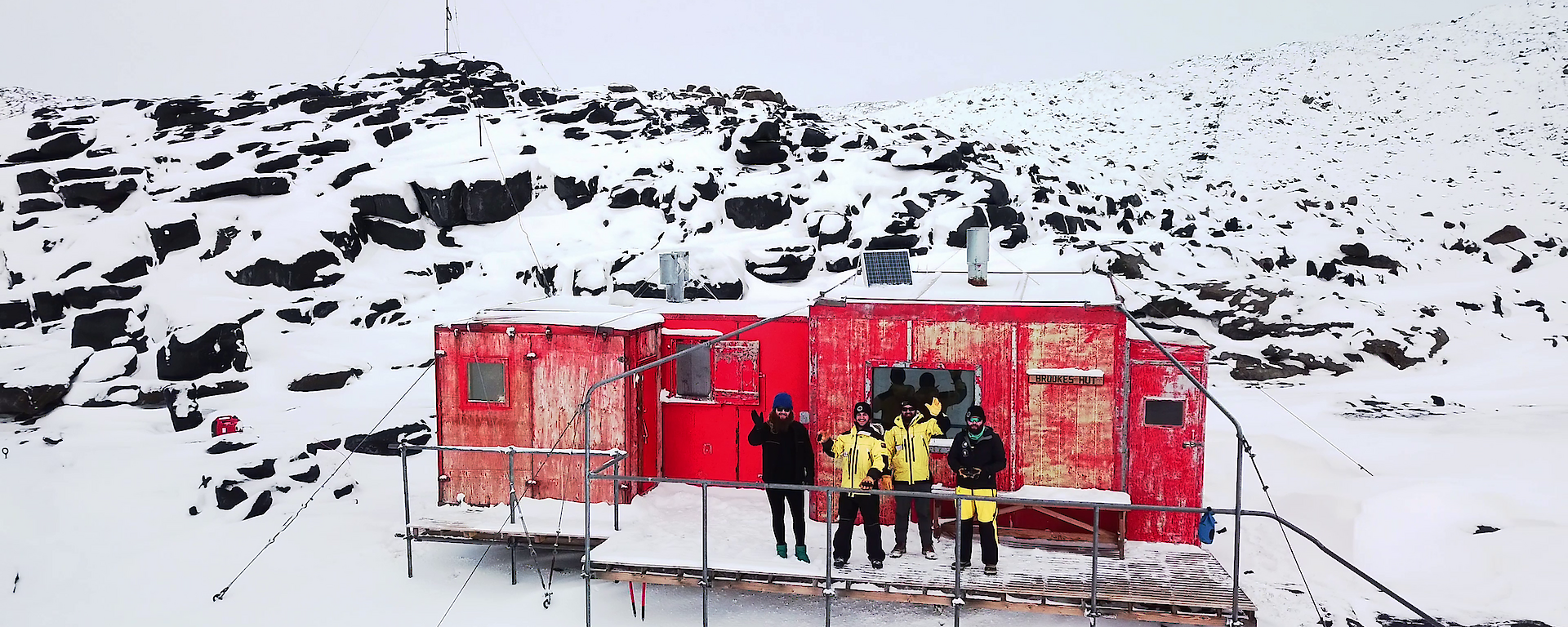 Aerial shot of four expeditioners standing on the veranda of a red hut waving to camera.  Surrounding landscape is snowy