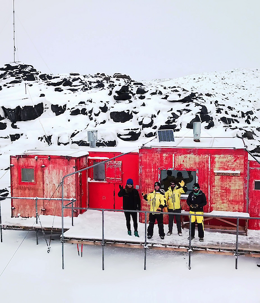 Aerial shot of four expeditioners standing on the veranda of a red hut waving to camera.  Surrounding landscape is snowy