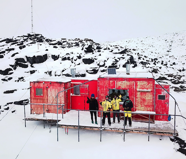 Aerial shot of four expeditioners standing on the veranda of a red hut waving to camera.  Surrounding landscape is snowy