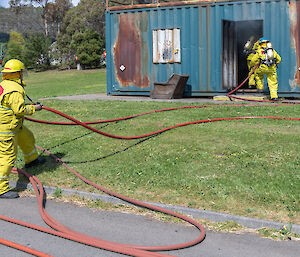 Firefighting students entering a smoke-filled building while another watches, holding the hose