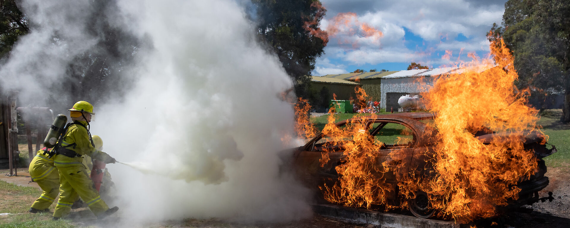 people in protective clothing use a fire extinguisher on a burning vehicle