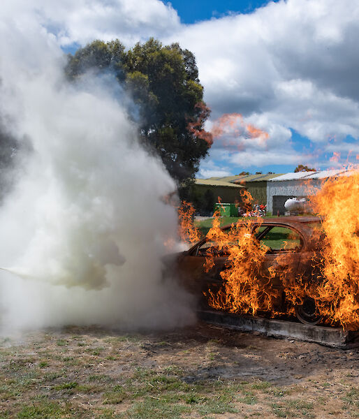 people in protective clothing use a fire extinguisher on a burning vehicle
