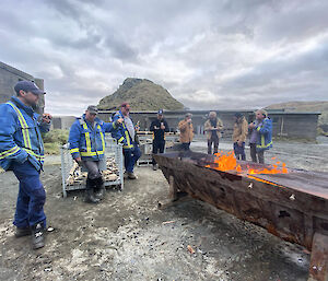 Group of guys standing around a metal container with a bonfire in it