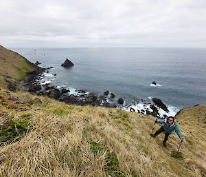 View looking down a tussocky hill to the ocean and rocks below with Alana standing on the hill further down