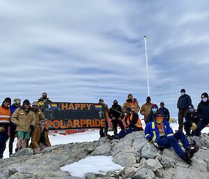 A group of expeditioners, outside the station, holding up a large flag with wording Happy Polar Pride