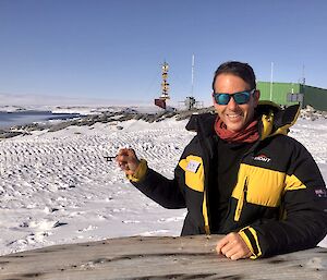 Station leader sits at an external wooden table with the station and anatarctic landscape in the background holding up a small key