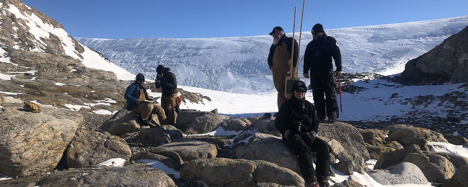 5 expeditioners stand and sit on some rocks in amongst the snow.  Snowy hills in the background