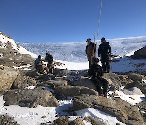 5 expeditioners stand and sit on some rocks in amongst the snow.  Snowy hills in the background