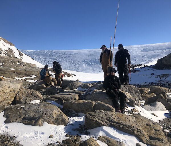 5 expeditioners stand and sit on some rocks in amongst the snow. Snowy hills in the background