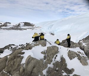 4 expeditioners sit on some snowy rocks looking out across a snowy landscape