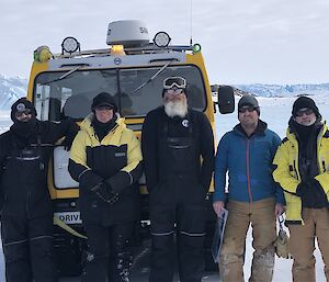 5 men standing smiling to camera in front of a yellow Hagglund