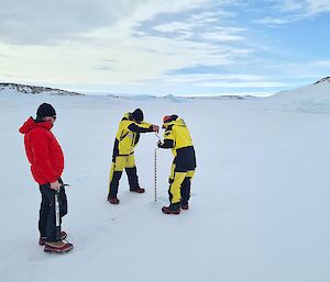 Two expeditioners operate a drill into the sea ice, supervised by a Field Training Officer.