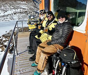 Three smiling expeditioners sitting on the veranda of the hut with cold beers