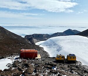 Two Hagglunds and a red hut on the rocky ground with snow patches