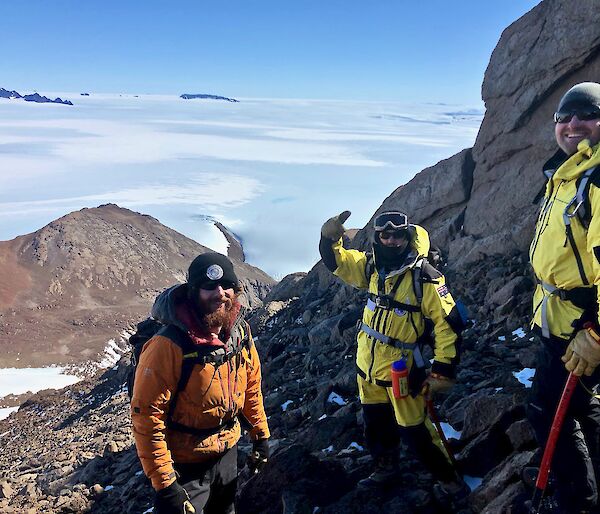 Three expeditioners smiling to camera near the summit of the mountain. Glacier and snow below