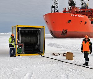 Brad standing beside a shipping container with a hose reel spooling out, with the Aurora Australis in the background and another person looking on.