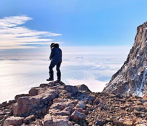 An Expeditiner standing on a rocky outcrop, looking as if he is about to step off.  The sea ice is visible far below.