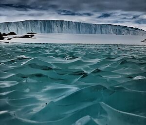 A jade coloured rippled pond that is frozen in the foreground.  A tabular iceberg in the background