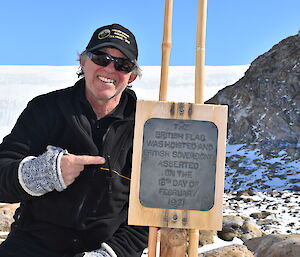 Expeditioner in baseball cap smiling to camera and pointing to Proclamation Point commemorative plaque