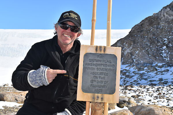 Expeditioner in baseball cap smiling to camera and pointing to Proclamation Point commemorative plaque