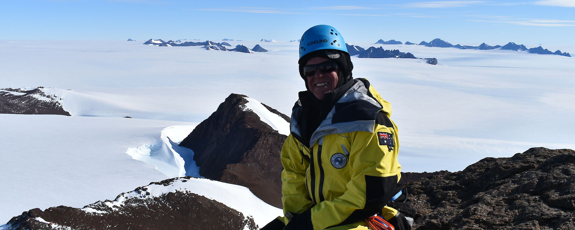 An expeditioner sitting on top of a mountain looking down towards a snow covered landscape