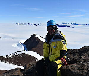 An expeditioner sitting on top of a mountain looking down towards a snow covered landscape