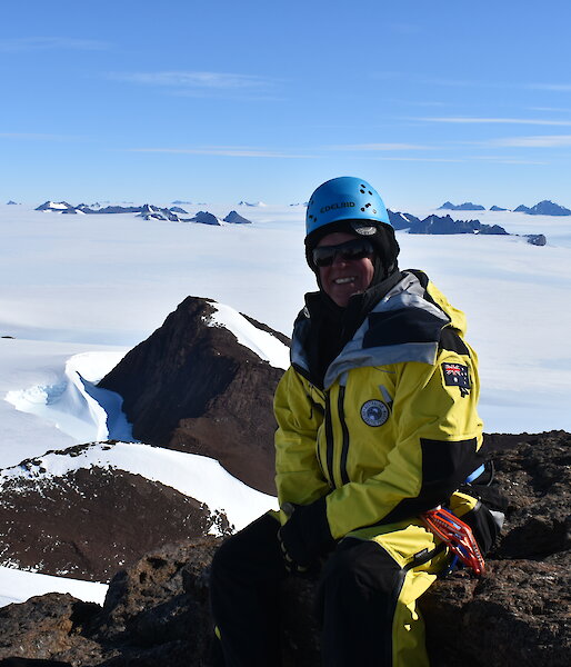 An expeditioner sitting on top of a mountain looking down towards a snow covered landscape