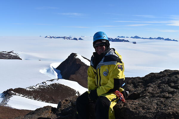 An expeditioner sitting on top of a mountain looking down towards a snow covered landscape