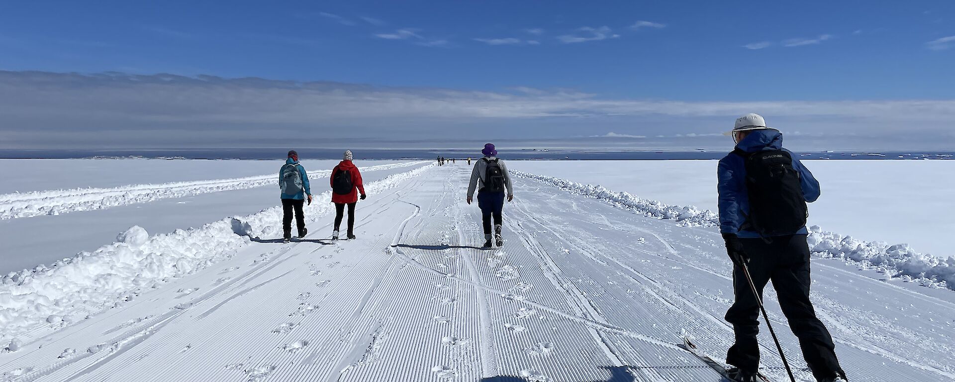 3 people walking. 1 person cross-country skiing. Groomed snow track. Blue skies. Ocean in the background.