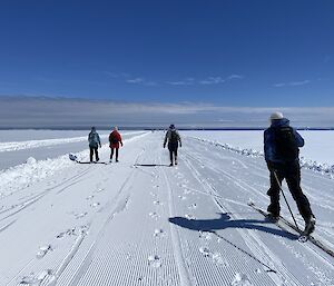 3 people walking. 1 person cross-country skiing. Groomed snow track. Blue skies. Ocean in the background.