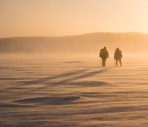 Two expeditioners walking to camera.  Snow is blowing and the sun is setting creating an orange glow