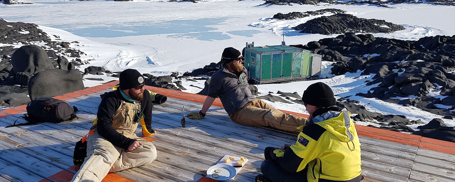 Three expeditioners sit relaxing on the wooden verandah of the hut with cheese and buscuits