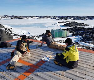 Three expeditioners sit relaxing on the wooden verandah of the hut with cheese and buscuits